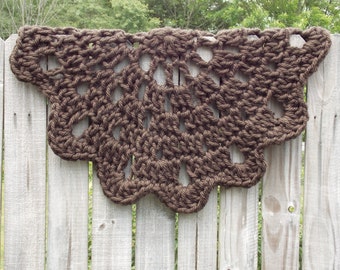May include: A brown, crocheted shawl with a scalloped edge hangs on a wooden fence. The openwork design of the shawl is visible, with a repeating pattern of diamond shapes. The yarn appears to be a thick, textured material.