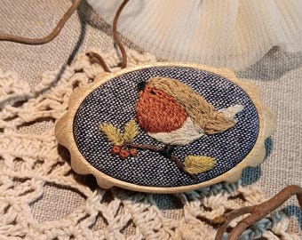 May include: An oval wooden brooch with an embroidered robin design. The bird has a red breast and tan head, perched on a branch with berries. The brooch is set against a blue and white tweed fabric.