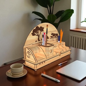 Puede incluir: Un organizador de escritorio de madera con tem&aacute;tica de la naturaleza. El organizador presenta un dise&ntilde;o de silueta de &aacute;rbol y elefante, con compartimentos para bol&iacute;grafos, l&aacute;pices y otros &uacute;tiles de oficina. Est&aacute; hecho de madera clara y tiene un aspecto c&aacute;lido y natural.