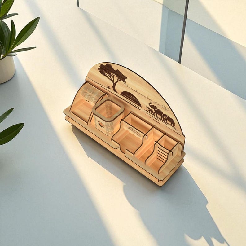 Puede incluir: Un organizador de escritorio de madera clara con m&uacute;ltiples compartimentos. Presenta un dise&ntilde;o cortado con l&aacute;ser de un &aacute;rbol, elefantes y el texto "We are the guardians of wildlife". Dise&ntilde;ado para guardar bol&iacute;grafos, l&aacute;pices y otros &uacute;tiles de oficina.
