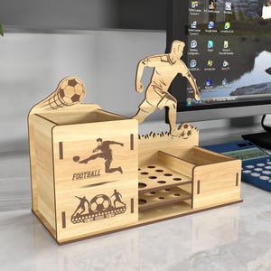 May include: Wooden desk organizer with a football theme. It features a cutout of a soccer player kicking a ball, a soccer ball, and the word "FOOTBALL". The organizer has a pen holder, a small drawer, and a section with multiple slots for holding pens or pencils.