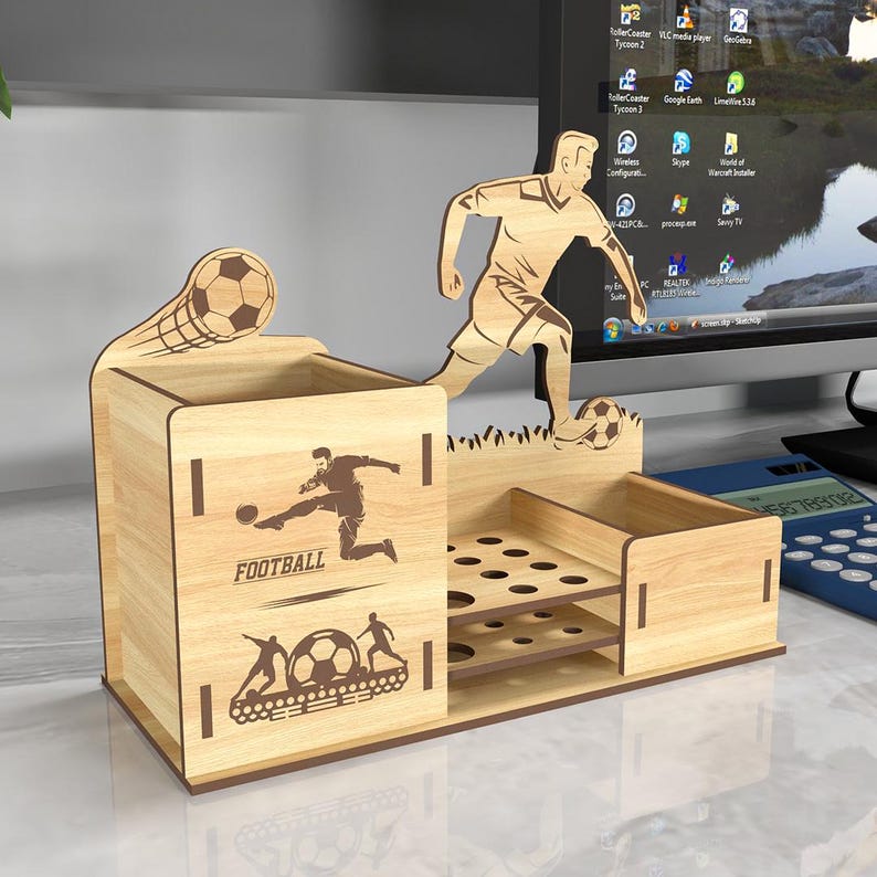 May include: Wooden desk organizer with a football theme. It features a cutout of a soccer player kicking a ball, a soccer ball, and the word "FOOTBALL". The organizer has a pen holder, a small drawer, and a section with multiple slots for holding pens or pencils.