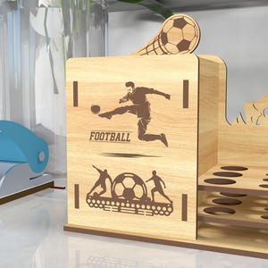 May include: Wooden desk organizer with a football theme. The organizer has a compartment for pens and pencils, a shelf for small items, and a cutout of a soccer player kicking a ball. The text "FOOTBALL" is printed on the organizer.