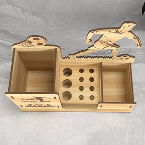 May include: Wooden desk organizer with a soccer player design. It has a large compartment, a smaller compartment, and a section with nine holes for pens or pencils. The words "FOOTBALL" are printed on the front.