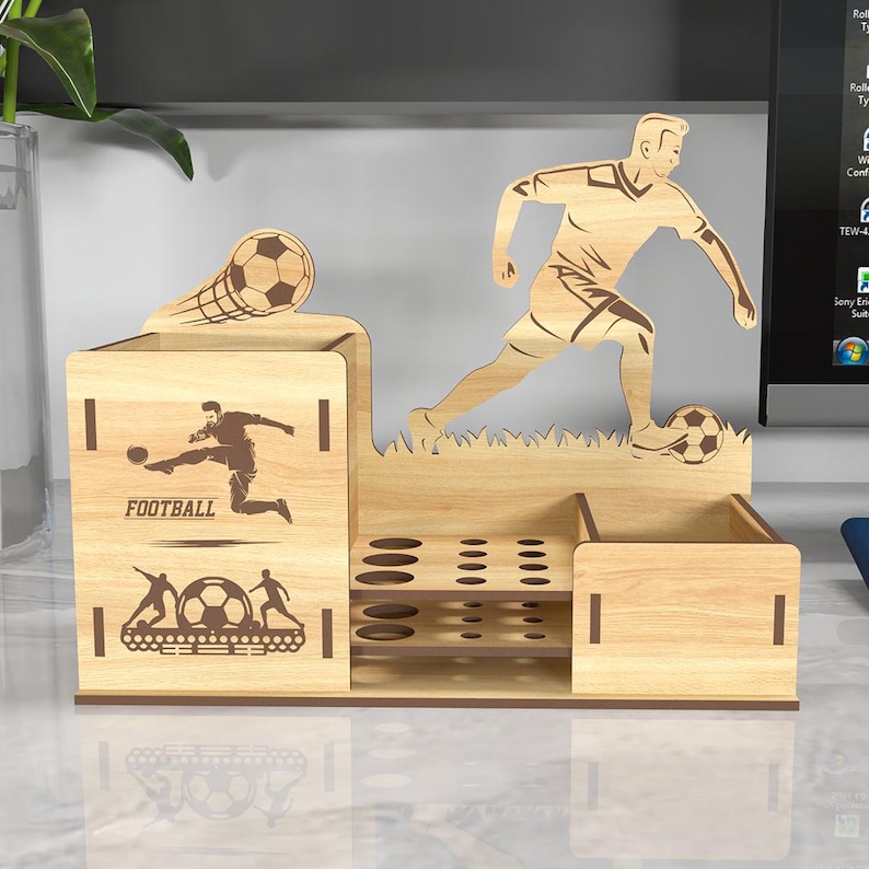 May include: Wooden desk organizer with a soccer theme. It features a silhouette of a soccer player kicking a ball, a soccer ball in a net, and the word "FOOTBALL". The organizer has multiple compartments for pens, pencils, and other office supplies.