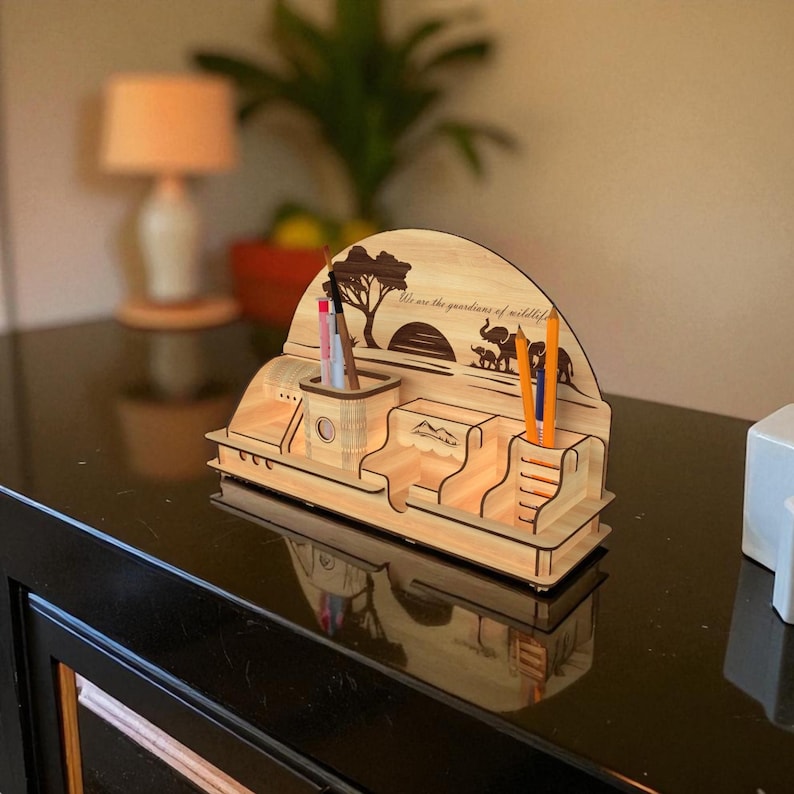 Puede incluir: Un organizador de escritorio de madera con tem&aacute;tica de vida silvestre. Cuenta con compartimentos para bol&iacute;grafos y objetos peque&ntilde;os, con un dise&ntilde;o cortado con l&aacute;ser de un &aacute;rbol, elefantes y el texto "We are the guardians of wildlife."