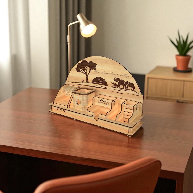 Puede incluir: Organizador de escritorio de madera con dise&ntilde;o tem&aacute;tico de la naturaleza. Presenta un &aacute;rbol, elefantes y el texto "We are the guardians of wildlife". Incluye compartimentos para bol&iacute;grafos, objetos peque&ntilde;os y un tel&eacute;fono. Madera clara.