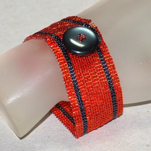 Op de afbeelding: Een geweven armband met zwarte en oranje strepen en een zwarte knoopsluiting.