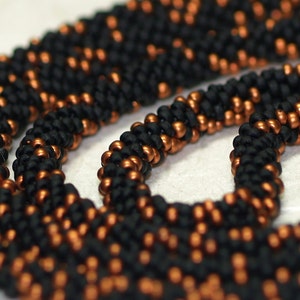 Puede incluir: Primer plano de un collar de cuentas negras y cobre. Las cuentas son pequeñas y están estrechamente entrelazadas, creando un diseño texturizado e intrincado.