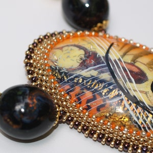 Op de afbeelding: Een close-up van een ketting met een grote, ovale hanger. De hanger is gemaakt van glas en heeft een kleurrijk abstract ontwerp met gouden accenten. De hanger is omgeven door een rand van kleine, gouden en bruine kralen.