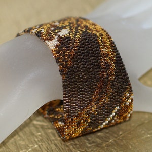 May include: A brown, gold, and white beaded bracelet with a geometric pattern. The bracelet is made of small, tightly woven beads.