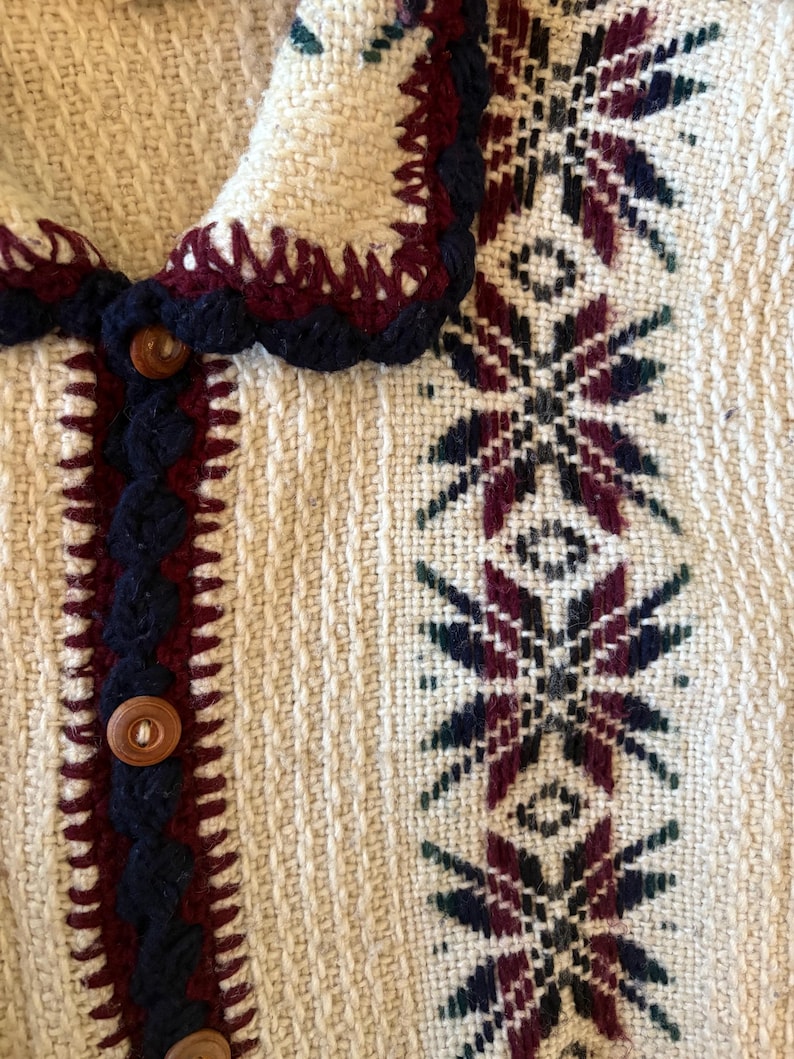 Puede incluir: Primer plano de una chaqueta tejida de color crema con un patr&oacute;n detallado. La chaqueta presenta un borde decorativo en azul oscuro y burdeos, con botones de madera y un dise&ntilde;o estampado en burdeos, azul oscuro y verde.
