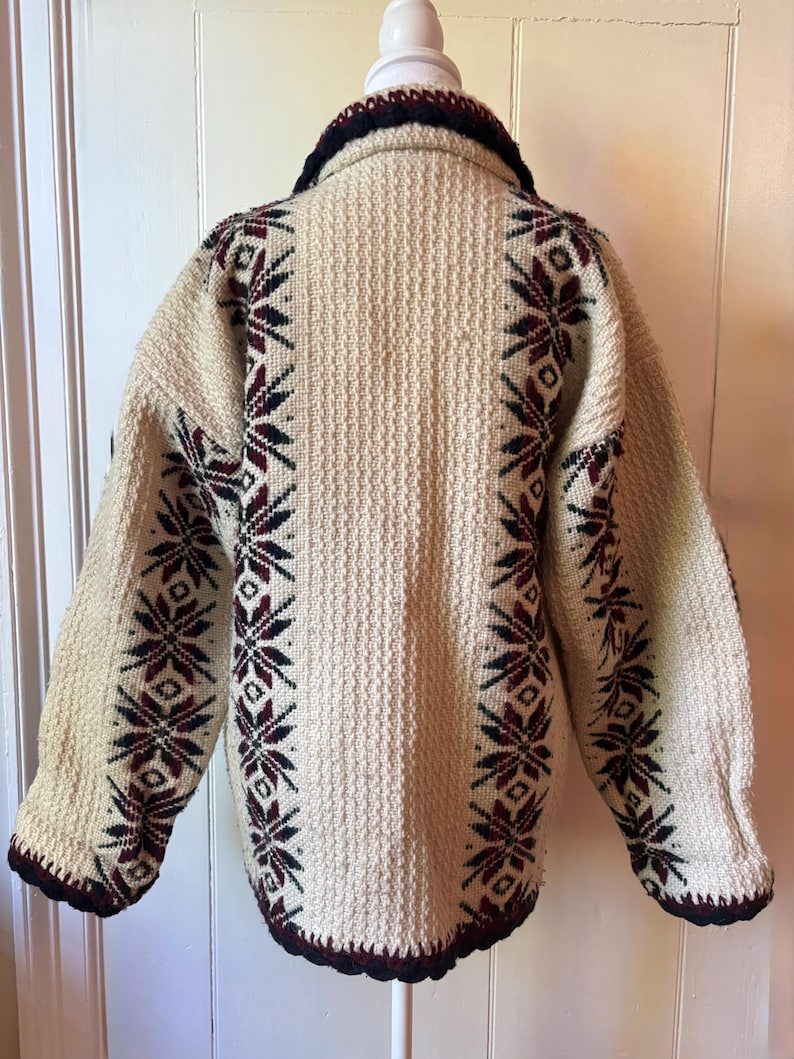 Puede incluir: Chaqueta de punto vintage color crema con tejido texturizado. Presenta cuello y ribetes azul marino oscuro, y patrones de copos de nieve en burdeos y azul marino a lo largo de las mangas y los laterales. Una prenda de abrigo c&aacute;lida y acogedora.