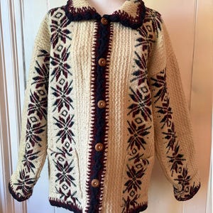 Puede incluir: C&aacute;rdigan de punto color crema con un estampado de copos de nieve en burdeos y azul marino. El su&eacute;ter tiene cuello, botones de madera y dos bolsillos delanteros. Los pu&ntilde;os y el cuello est&aacute;n ribeteados en azul marino.