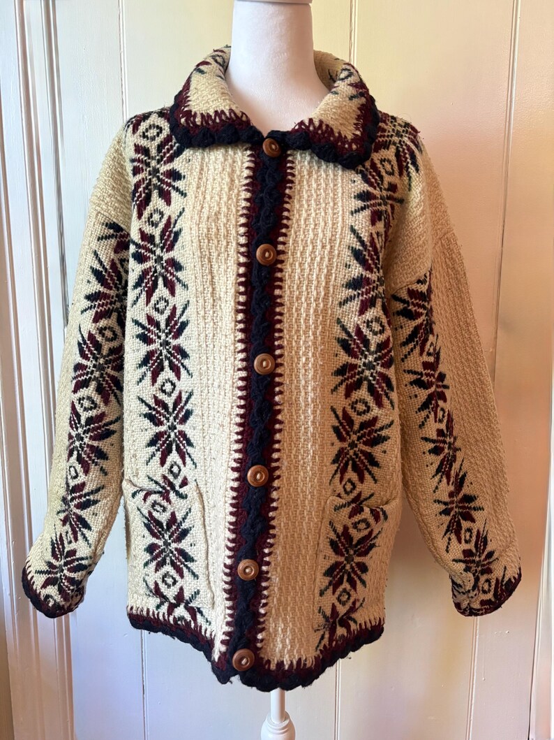 Puede incluir: C&aacute;rdigan de punto color crema con un estampado de copos de nieve en burdeos y azul marino. El su&eacute;ter tiene cuello, botones de madera y dos bolsillos delanteros. Los pu&ntilde;os y el cuello est&aacute;n ribeteados en azul marino.