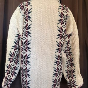Puede incluir: Chaqueta tejida de color crema con cuello y ribete azul marino. La chaqueta presenta un patr&oacute;n repetido de dise&ntilde;os de copos de nieve burdeos y azul marino a lo largo de las mangas y los lados. La chaqueta tiene una apariencia texturizada.