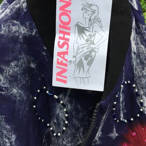 Puede incluir: Una chaqueta de color morado y blanco con estampado tie-dye, con una cremallera negra y una etiqueta blanca que dice "INFASHIONS". La chaqueta tiene un dise&ntilde;o de cuentas en la parte delantera.