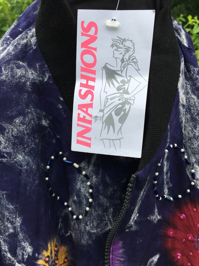 Puede incluir: Una chaqueta de color morado y blanco con estampado tie-dye, con una cremallera negra y una etiqueta blanca que dice "INFASHIONS". La chaqueta tiene un dise&ntilde;o de cuentas en la parte delantera.