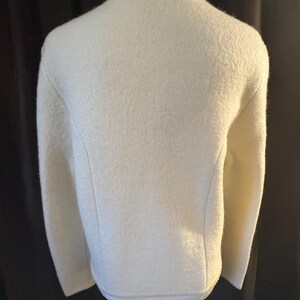 May include: Cream-colored wool jacket with a textured surface. The jacket has a round neckline and long sleeves. The back view shows a tailored fit with subtle seams and a decorative trim along the neckline and hem.