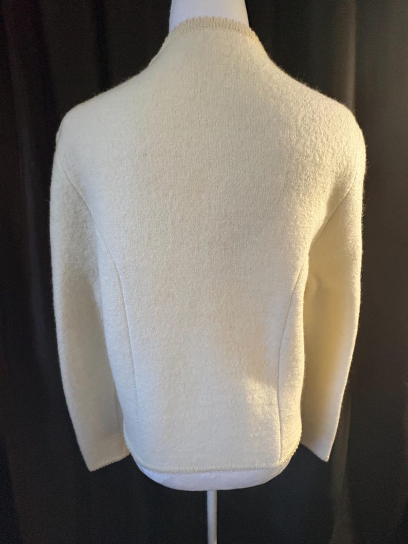 May include: Cream-colored wool jacket with a textured surface. The jacket has a round neckline and long sleeves. The back view shows a tailored fit with subtle seams and a decorative trim along the neckline and hem.
