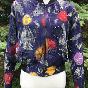 Puede incluir: Una chaqueta bomber de color morado, blanco y rojo con estampado tie-dye y adornos de cuentas. La chaqueta tiene cierre de cremallera y cintura de canal&eacute;.