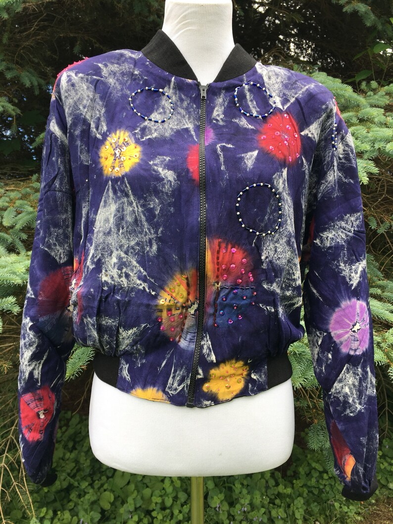 Puede incluir: Una chaqueta bomber de color morado, blanco y rojo con estampado tie-dye y adornos de cuentas. La chaqueta tiene cierre de cremallera y cintura de canal&eacute;.