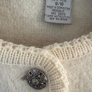 May include: Close-up of a cream-colored wool cardigan with a decorative silver button. The label reads "100% Wool" and "Made in Dominican Republic". The cardigan has a textured knit design.