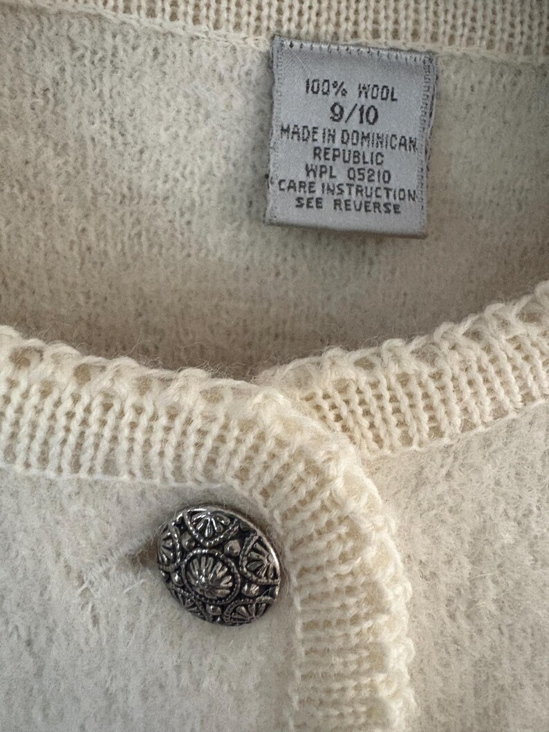 May include: Close-up of a cream-colored wool cardigan with a decorative silver button. The label reads "100% Wool" and "Made in Dominican Republic". The cardigan has a textured knit design.