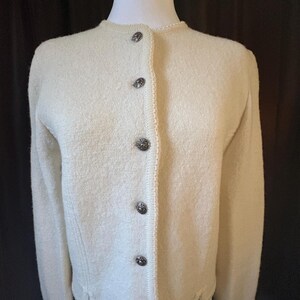 May include: Cream-colored cardigan sweater with a round neckline and long sleeves. The sweater features decorative buttons down the front and a textured knit design. The buttons are dark with a decorative pattern.