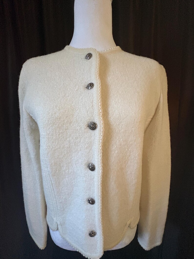 May include: Cream-colored cardigan sweater with a round neckline and long sleeves. The sweater features decorative buttons down the front and a textured knit design. The buttons are dark with a decorative pattern.