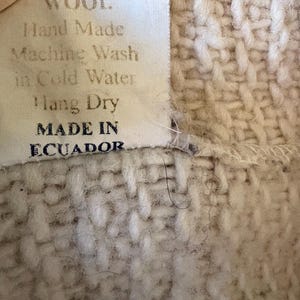 Puede incluir: Primer plano de un textil tejido con una etiqueta. La etiqueta dice "WOOL. Hand Made Machine Wash in Cold Water Hang Dry MADE IN ECUADOR." El textil es de color beige claro con un patr&oacute;n tejido texturizado.