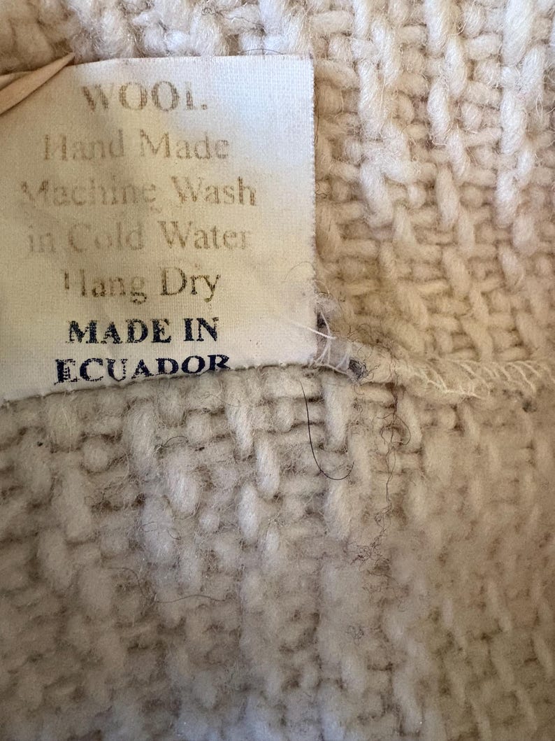 Puede incluir: Primer plano de un textil tejido con una etiqueta. La etiqueta dice "WOOL. Hand Made Machine Wash in Cold Water Hang Dry MADE IN ECUADOR." El textil es de color beige claro con un patr&oacute;n tejido texturizado.