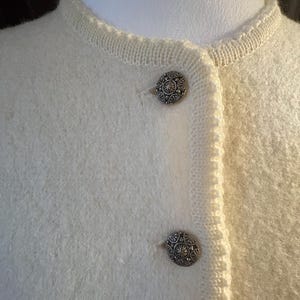 May include: Close-up of a cream-colored cardigan with two ornate silver buttons. The cardigan has a textured, fuzzy appearance and a delicate knit trim around the neckline and button placket. The buttons are detailed with a floral design.