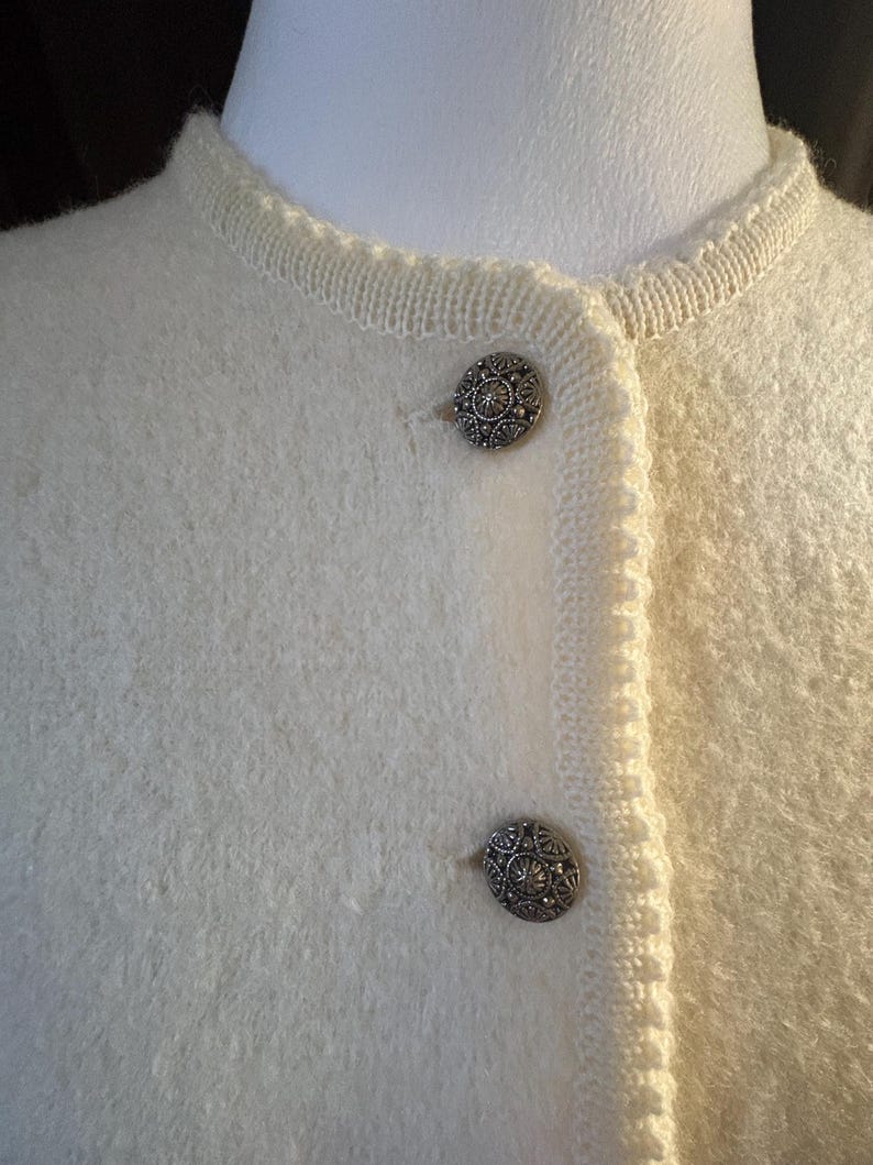 May include: Close-up of a cream-colored cardigan with two ornate silver buttons. The cardigan has a textured, fuzzy appearance and a delicate knit trim around the neckline and button placket. The buttons are detailed with a floral design.