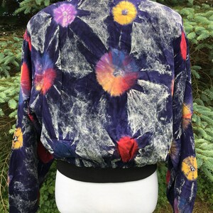Puede incluir: Una chaqueta bomber azul con un estampado tie-dye con flores de colores. La chaqueta tiene un cuello, pu&ntilde;os y cintura de canal&eacute; negros.