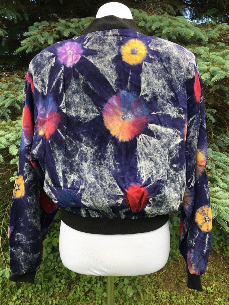 Puede incluir: Una chaqueta bomber azul con un estampado tie-dye con flores de colores. La chaqueta tiene un cuello, pu&ntilde;os y cintura de canal&eacute; negros.