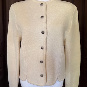 May include: Cream-colored cardigan with a round neckline and long sleeves. The button-down sweater features decorative stitching and dark brown buttons. The hemline has a scalloped edge. A classic, vintage style.