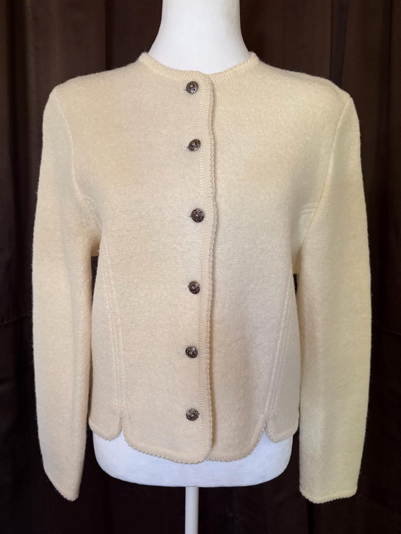 May include: Cream-colored cardigan with a round neckline and long sleeves. The button-down sweater features decorative stitching and dark brown buttons. The hemline has a scalloped edge. A classic, vintage style.