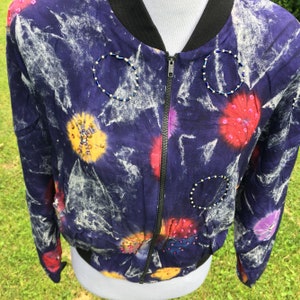 Puede incluir: Una chaqueta bomber azul con un estampado tie-dye y adornos de cuentas de colores. La chaqueta tiene una cremallera y un cuello negro.