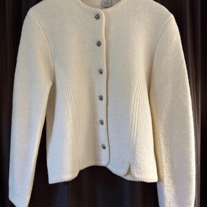 May include: Off-white wool jacket with a round neckline and long sleeves. The jacket has decorative stitching and is fastened with six silver-toned buttons. The hemline and neckline are trimmed with a delicate, textured border.