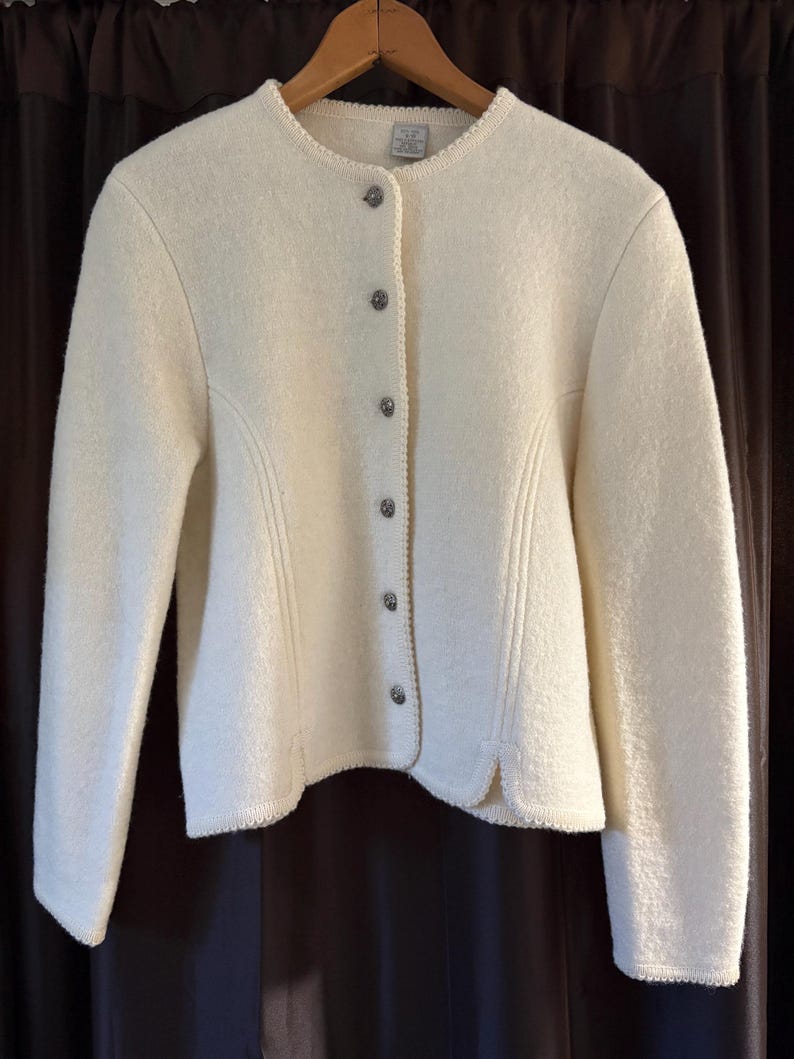 May include: Off-white wool jacket with a round neckline and long sleeves. The jacket has decorative stitching and is fastened with six silver-toned buttons. The hemline and neckline are trimmed with a delicate, textured border.
