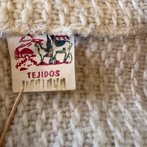 Puede incluir: Primer plano de una tela de punto color crema con una etiqueta rectangular. La etiqueta presenta una ilustraci&oacute;n roja y azul de animales y la palabra "TEJIDOS". La tela tiene un aspecto texturizado y tejido.