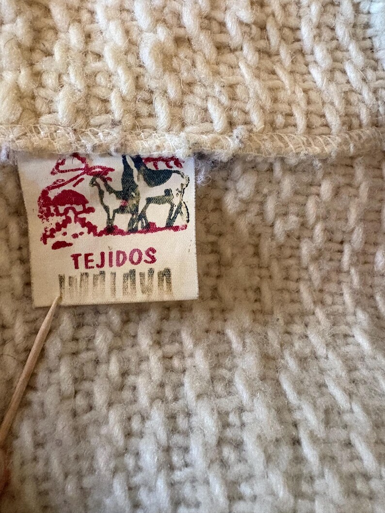 Puede incluir: Primer plano de una tela de punto color crema con una etiqueta rectangular. La etiqueta presenta una ilustraci&oacute;n roja y azul de animales y la palabra "TEJIDOS". La tela tiene un aspecto texturizado y tejido.