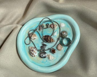 May include: An irregular, light blue, glossy trinket dish containing a silver chain necklace with a rectangular pendant, a separate silver chain, a silver ring, and several small, natural, multi-coloured seashells scattered across the surface.