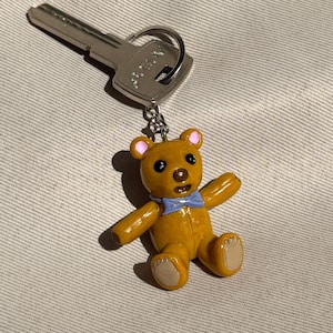 Puede incluir: Un llavero con un pequeño dije de oso de peluche amarillo. El oso tiene una pajarita azul, orejas rosas y patas color canela. Una llave plateada está unida al llavero.