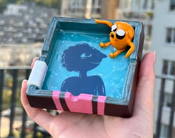 May include: A square ashtray with a dark brown exterior and a bright blue interior. The centre features a dark blue silhouette of a figure. A small, orange, cartoon-style dog character is attached to the rim. The rim has pink and black stripes.