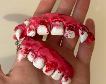 May include: A keyring featuring two sculpted, realistic-looking gum lines with white teeth. The gums are painted a vibrant pink and are splattered with dark red paint to resemble blood. The teeth are glossy, white, and include pointed fangs.