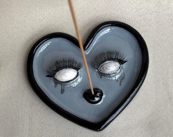May include: A heart-shaped incense holder in glossy grey with a thick black rim. The surface features two sculpted eyes with long black lashes and teardrop details, one with a white iris and one with a black and white swirl, plus a black incense stick.
