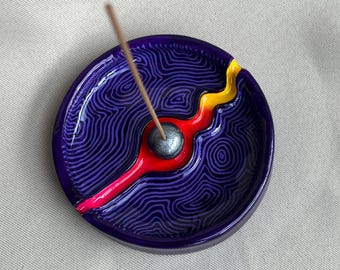 May include: A circular ceramic incense holder featuring a deep purple base with intricate, swirling black topographic line patterns. A vibrant red and yellow channel cuts across the centre, holding a metallic grey sphere with an incense stick.