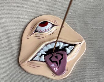 May include: A beige ceramic incense holder shaped like a face with a single red eye and an open mouth showing white teeth. A purple tongue protrudes from the mouth, featuring a black geometric symbol. A thin brown incense stick is inserted.
